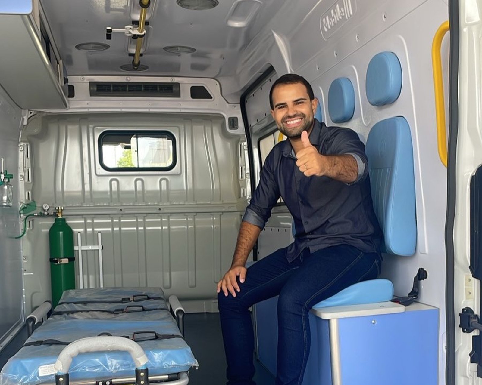 Guilherme Eduardo sentado dentro de uma ambulância, fazendo sinal de positivo, com a maca e os equipamentos ao lado