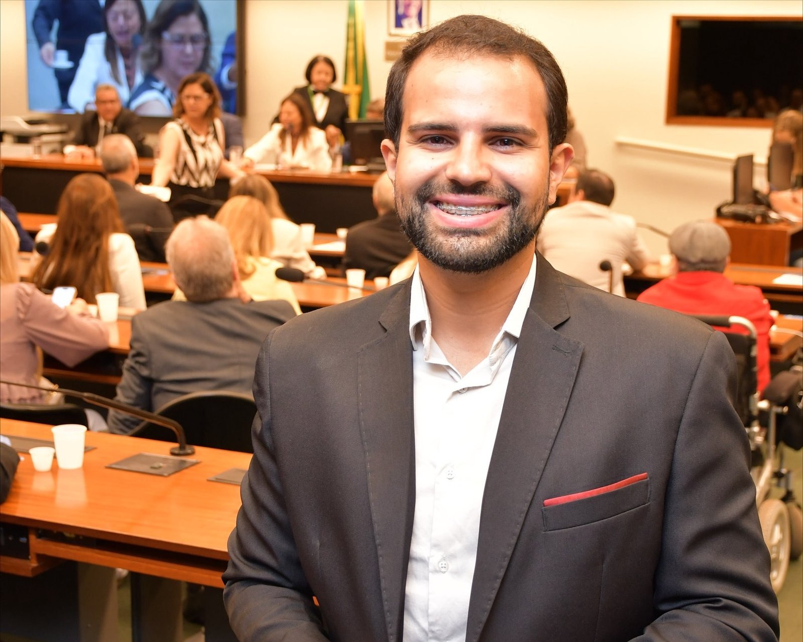 Guilherme Eduardo de terno, sorrindo em um auditório lotado durante um evento institucional